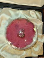 Top-down view of round glass paperweight showing red interior with central circular glass element and smaller air bubbles. Displayed in satin-lined box.