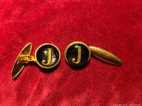 Pair of gold tone cuff links with black enamel face featuring a gold letter 'J'. Displayed on red velvet background showing front and side profiles.