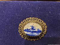 Close-up photo of the brooch showing detailed gold filigree frame and the blue and white windmill painting on porcelain cabochon.