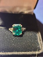 Close-up photo of silver tone ring with emerald cut green stone in black velvet ring box.