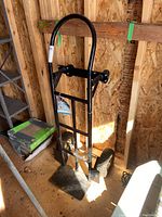 Full view of Ultra Steel black steel heavy duty hand truck dolly standing upright in a wooden shed, with its curved handle, base plate, and pneumatic wheels visible.