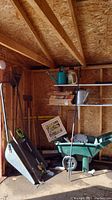 Wide view of shed corner showing various garden tools including rakes, shovel, watering cans, broom, and wheelbarrow.