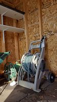 Photo showing portable resin hose reel with green garden hose and spray nozzle, placed inside a wooden shed. Includes close-up of the reel and hose coiled around it.