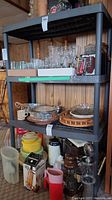 Full view of black resin shelving unit displaying 4 shelves loaded with glassware, plastic containers, and kitchen items. Shelving unit in a room with wood-paneled walls.
