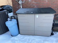 Outdoor Rubbermaid storage shed with black lid and beige body next to a brick wall, snow on ground, shed doors closed