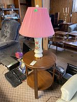 Side view of round wooden side table with lower shelf and ceramic table lamp with pink pleated shade on top.