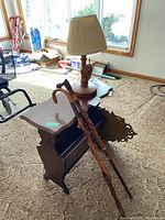 Side and front angle of wooden magazine rack table with two carved wooden walking canes leaning on it and wood-based lamp on top.