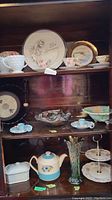Shelf showing various teacups, saucers, carnival glass vase, ruffled bowl, floral decorative plates, hobnail creamer, and sugar.