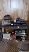 Wide photo of all the electronics and accessories including turntable, media players, speakers, cables, and tapes on a plastic table with wood panel wall background.