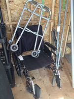 Four-wheeled height adjustable walker with no brakes shown leaning on a Jennings wheelchair inside a wooden paneled storage area.