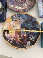 Circular porcelain wall mural with colorful depiction of girl, cat, and flowers. Measuring tape showing 14 inch diameter.
