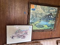 Two framed artworks displayed on a wood-paneled wall above a striped sofa, one with mountain lake scene and the other a harbor scene.