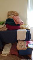 Stack of assorted bedspreads, linens, and sheets on table near window showing various folded fabric items in solid colors and patterns.