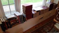 Wide view showing collection of books placed by a window, including sets of thick hardcover volumes with red and blue spines, various sized books stacked and lined up