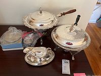 Photo of two silver plated chafing dishes, footed bowl, glass bowl with metal rim and fork, and gravy boat displayed together on wooden surface.