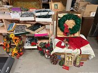 Photo showing multiple Christmas decorations on the floor including a large green wreath with red ribbon, smaller wreaths with autumn leaves and berries, boxes of string lights, silver tinsel, pillar candles, and assorted decorations.