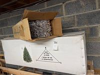 Long white cardboard box labeled “7ft Botoner Pine Tree” with green tree illustration and triangle diagram, topped by smaller open brown box of branches.