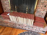 Full view of 24 bound volumes arranged on a fireplace hearth showing red leather or faux leather covers with gold embossed decoration and titles.