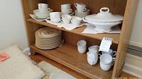 Photo showing shelves with stacked plates, cups, and milk glass tea cups along with covered serving dish on display