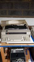 IBM Wheelwriter 3 electric typewriter placed on a wooden shelf against a concrete block wall, showing the front view with keyboard and display panel, accompanied by a power cord and some papers placed on top.