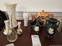 Overview of Lenox porcelain vases, pottery green handled pieces with classical relief, and a ceramic cottage teapot on wood surface