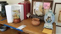 Overview table photo showing a nativity snow globe in Styrofoam packaging, plastic model ship, small woven basket, and cherub music box alongside framed background decorations.