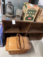 Wicker picnic basket on shelf with two metal lanterns and boxed Coleman heater behind