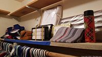 Shelf with multiple boxed formal handkerchiefs in white packaging, NASCAR caps stacked on top of each other, a striped fabric pouch, and a plaid patterned thermos bottle.