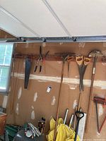 Wall-mounted handsaws, pruning shears, metal rake and garden tools hung on wood panel in garage.
