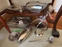Silverplate serving trays, wire basket casserole with lid, cup, and silver barware pieces on floor under table.