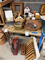 Photo showing assorted kitchen and dining items including baskets, wooden trays, mugs, a metallic gold bowl, and other accessories arranged on and under a table.