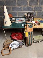 Photo showing a variety of vintage sporting goods including a golf bag with clubs, wooden tennis rackets with Spalding covers, bowling ball and shoes, basketball, megaphone, leather bag, gloves, and other small items on a table and floor.