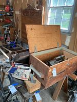 Two wooden tool crates in a garage workshop setting. The open crate contains a Dremel Moto Jig Saw, various hardware, and tool manuals. The second crate holds a metal vise, drum sander sleeves, several jack planes, and miscellaneous hand tools. The crates and tools show wear consistent with heavy use.