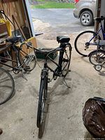 Front angled view of the vintage black Triumph bicycle stored inside a garage, showing the full bike frame, handlebars, seat, and front wheel.