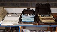Photo showing three electric typewriters on shelf, cases open and dust visible, with one white Brother, one blue unbranded, and one beige Smith-Corona Super 12.