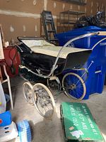 Front-side view showing black outer shell, cream interior lining, and cream sun blocker canopy on a metal frame with spoked wheels.