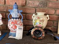 Both urns shown together on a shelf in front of a brick wall. One urn has a blue lid and dragon-shaped handles with orange pomegranate decoration. The other is a beige urn with floral and butterfly motif and two small handles.