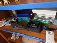 Shelf with two vintage metal staplers, Canon digital camera in black case, and Lifelong sealed gift watches in colorful packaging stacked behind.