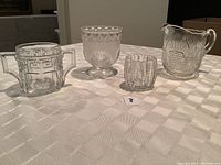 Four antique pressed glass pieces of varying sizes ranging from approximately 2 1/2 to 5 inches displayed on a white tablecloth.