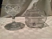 Two antique pressed glass items on a table with white textured cloth: a compote and a covered dish with handle and lid.