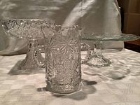 Photo of four antique pressed glass items including compote, celery vase, flat cake plate and flower shaped serving plate on white cloth backdrop.