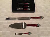 Four Christmas-themed serving utensils with decorative red, green, and silver handles laid out on white tablecloth.