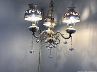 Chandelier lit up with three lights on, showing glowing perforated metal shades and hanging ceramic flowers.