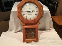 Front view of oak chiming wall clock showing the octagonal shape, Roman numeral face, and lower glass pendulum door.