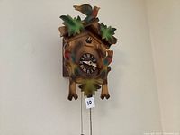 Front view of wooden cuckoo clock mounted on wall showing carved birds and leaves and clock face with Roman numerals