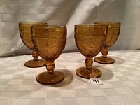 Four amber wine glasses shown from the front on a white cloth background with detailed floral embossing on the bowl and faceted short stem.