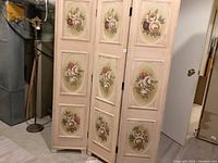 Full view of the 3-panel wooden room divider painted beige with floral artwork on each panel.