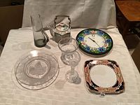 Overview of painted glass floral clock, ceramic tea pot, tall glass vase, cut glass bowl, and two plates including etched glass and floral porcelain.