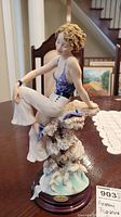 Full view of the porcelain figurine showing a seated woman with curly hair atop a textured rocky base on a wooden stand.