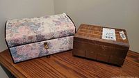 Two jewelry boxes on wooden surface: one tapestry fabric covered with floral pattern, one vintage wood box with latch.